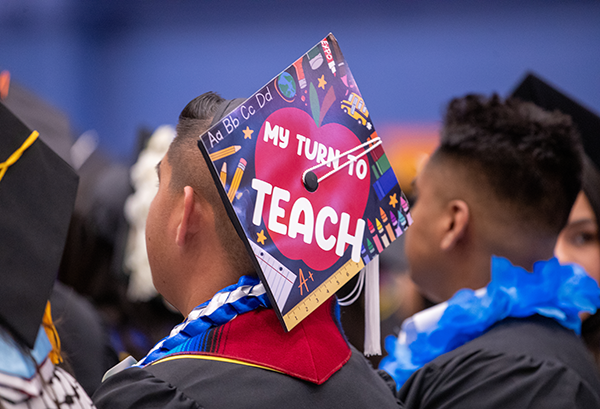 A close up on the back of a student sitting at a commencement ceremony with a graduation cap decorated with graphics of teacher and school supplies and a large apple in the middle of the hat with text over it reading "My turn to teach."