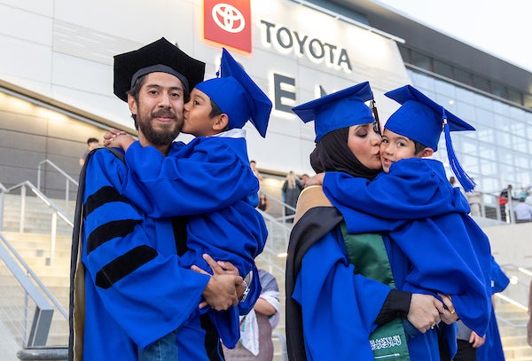 Two UCR graduate parents stand next to each other in cap and gown while each holding a young child.