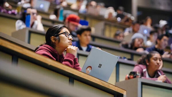 A student with glasses sits in the foreground of a lecture hall surrounded by other students behind and around her.