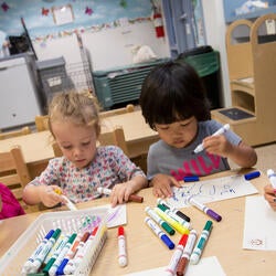 A group of 4 children sit at a table drawing pictures with colorful markers.