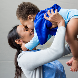 A smiling woman holds a smiling baby with their foreheads touching.