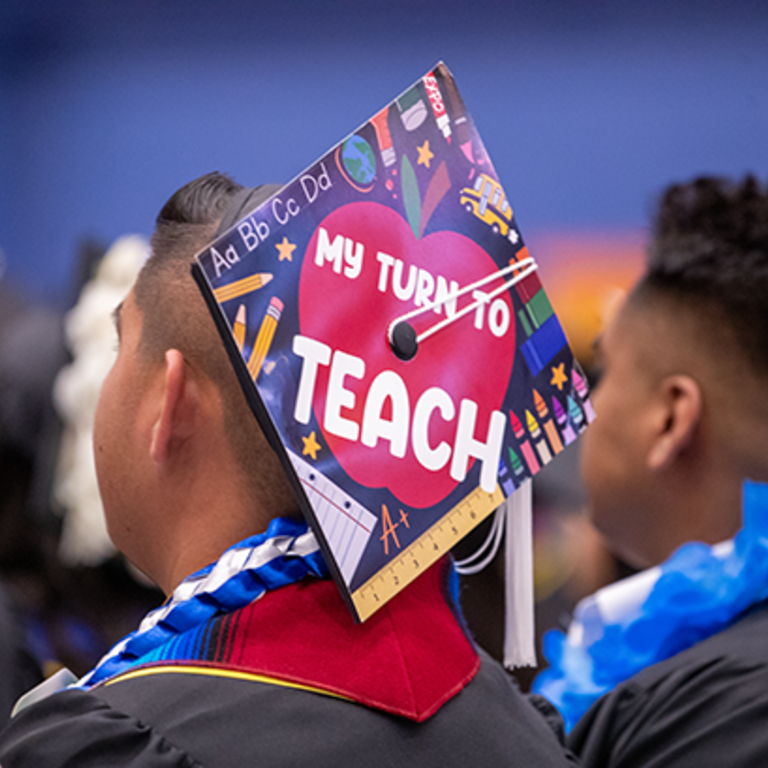 A close up on the back of a student sitting at a commencement ceremony with a graduation cap decorated with graphics of teacher and school supplies and a large apple in the middle of the hat with text over it reading "My turn to teach."