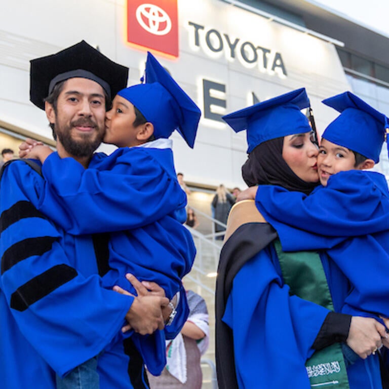 Two UCR graduate parents stand next to each other in cap and gown while each holding a young child.